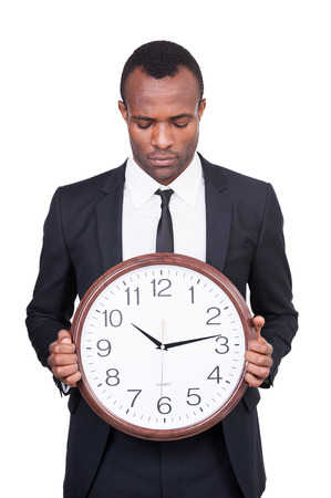 What time is it now? Serious young African man in formalwear holding a clock in his hand and looking at it while standing isolated on white background の写真素材