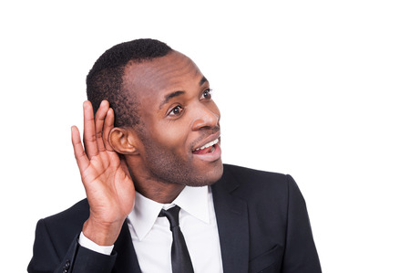 What did you say? Cheerful young African man in formalwear holding hand near ear and smiling while standing isolated on white background の写真素材