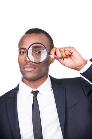 Businessman examining you. Serious young African man in formalwear looking through a magnifying glass while standing isolated on white background の写真素材