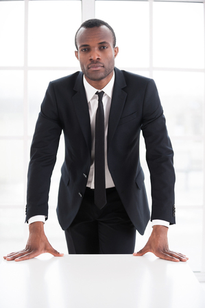 Confident and successful. Thoughtful young African man in formalwear leaning at the table and looking at cameraの写真素材