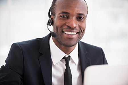Customer service representative. Cheerful young African man in formalwear and headset working on laptop and smiling at cameraの写真素材