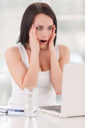 Shocked business lady. Surprised young woman holding head in hands and looking at the laptop while sitting at the tableの写真素材