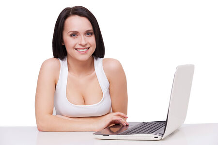 Beauty with laptop. Attractive young woman with beautiful cleavage working on laptop and smiling while sitting at the table and isolated on whiteの写真素材