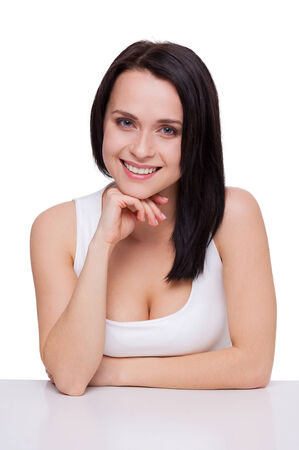 Cute and confident. Attractive young woman with beautiful cleavage gesturing and smiling while sitting at the table and isolated on whiteの写真素材