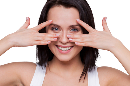 Look in my eyes! Attractive young woman holding hands near eyes and smiling while standing isolated on whiteの写真素材
