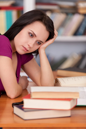 Tired student. Tired young woman holding his head in hand and looking at the book stack while sitting at the library deskの写真素材