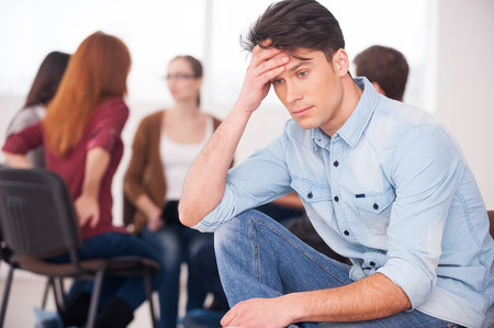 Feeling pain and depression. Depressed young man sitting at the chair and holding head in hand while other people communicating on background の写真素材