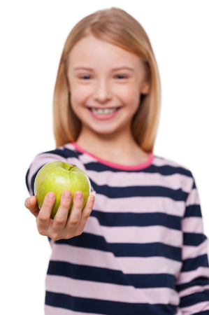 Sharing apple with you. Cheerful little girl stretching out hand with green apple while standing isolated on whiteの写真素材