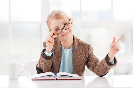 Look at that! Cheerful little girl in glasses and formalwear sitting at the table and pointing awayの写真素材