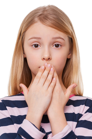 Oops! Frustrated little girl covering mouth with hands and looking at camera while standing isolated on whiteの写真素材