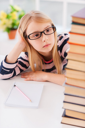 Tired of studying. Tired little girl looking at the book stack and holding head in hand while sitting at the tableの写真素材