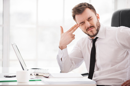 Depressed and frustrated. Depressed young man in formalwear touching his temple with finger gun and keeping eyes closed while sitting at his working placeの写真素材