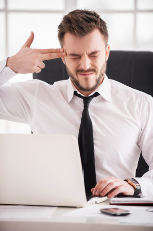 Monday morning suicide. Frustrated young man in formalwear touching his temple with finger gun and keeping eyes closed while sitting at his working placeの写真素材