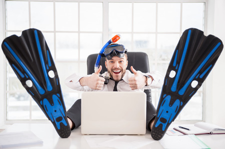 Ready for vacation. Handsome young office worker in in snorkel and flippers gesturing and smiling while sitting at his working placeの写真素材