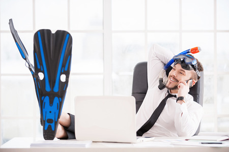 Booking tickets for vacation. Handsome young office worker in in snorkel and flippers gesturing and smiling while sitting at his working placeの写真素材