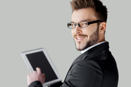 Confident in his tablet. Rear view of cheerful young man in formalwear and glasses working on digital tablet and looking over shoulder while standing against grey backgroundの写真素材