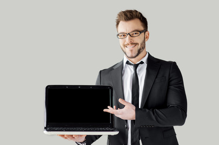 Pointing copy space on his laptop. Handsome young man in formalwear and glasses holding a laptop and pointing it while standing against grey backgroundの写真素材
