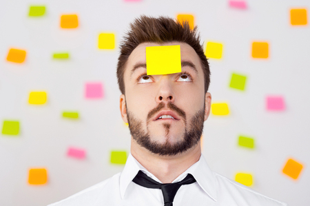 So much work to do! Portrait of frustrated young man in formalwear and adhesive note on his forehead standing against the wall with many adhesive notes on it の写真素材