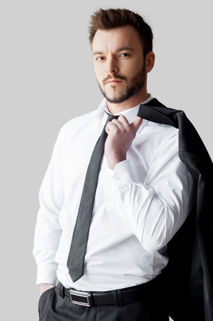 Relaxed businessman. Handsome young man in formalwear holding his jacket on finger and looking at camera while standing against grey backgroundの写真素材