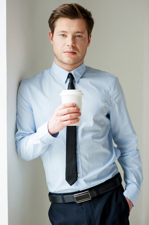 Taking a coffee break. Handsome young man in shirt and tie holding a cup of coffee and looking at cameraの写真素材