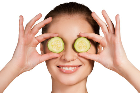 Cucumber spa. Beautiful young woman holding pieces of cucumber in front of her eyes and smiling while isolated on white backgroundの写真素材