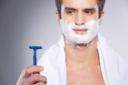 Young shirtless man looking at the razor and expressing negativity while standing isolated on grey backgroundの写真素材