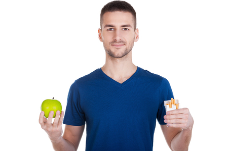 Make the right choice. Confident young man holding apple in one hand and pack of cigarettes in another one and isolated on whiteの写真素材
