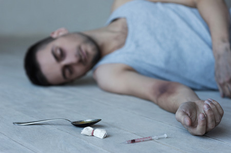 Heroin addict. Young man lying on the floor with syringe and pack of heroin laying near him の写真素材