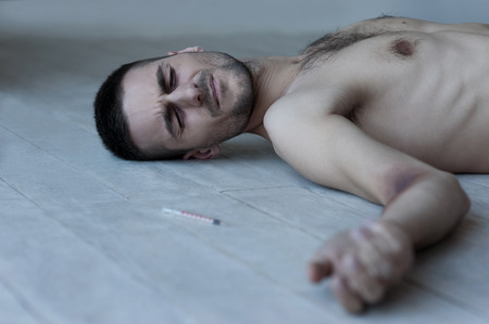  Young shirtless man lying on the floor and keeping eyes closed while syringe with narcotic laying near himの写真素材