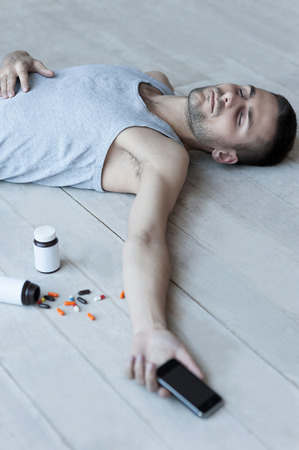  Top view of young man lying on the floor and holding a mobile phone in his hand while bottles with pills laying near himの写真素材