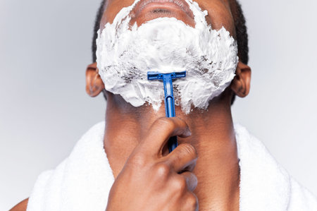Close up of man shaving. Close up of young African man shaving his face while standing against grey...
