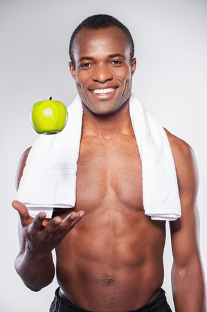 Enjoying healthy lifestyle. Young muscular African man with towel on shoulder throwing up an apple and smiling at camera while standing against grey backgroundの写真素材