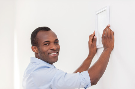 Man hanging picture frame. Cheerful young black man hanging picture frame and smiling at cameraの写真素材