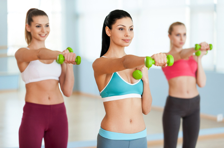 Exercising with dumbbells. Three beautiful young women in sports clothing exercising with dumbbells and smilingの写真素材