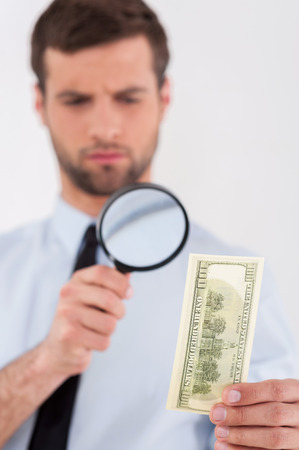 Fake or real? Concentrated young man in shirt and tie looking through a magnifying glass at the paper currency while standing isolated on white background の写真素材