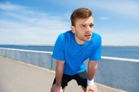 Tired of running. Tired young man holding hands on knees and looking away while standing outdoors