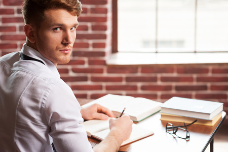 Confident author. Handsome young man in shirt and tie writing something in note pad and looking over shoulder while sitting at his working place の写真素材