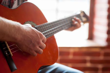 Playing acoustic guitar. Close-up of man playing acoustic guitar while sitting in front of the windowの写真素材