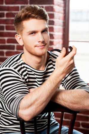 Handsome smoking pipe. Cheerful young man in striped shirt holding pipe and looking at camera while sitting on the chair の写真素材