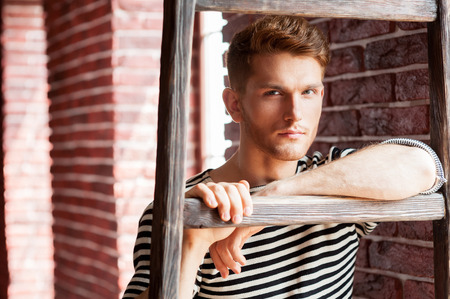Young and handsome. Handsome young man in striped shirt leaning at the wooden ladder and looking at camera while standing near the brick wallの写真素材