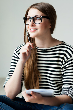 Just inspired. Thoughtful young woman in striped clothing holding note pad and touching her chin with pen while sitting on the hardwood floorの写真素材