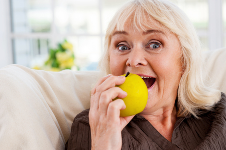 I love apples! Cheerful senior woman holding apple and looking at camera while sitting at the chairの写真素材