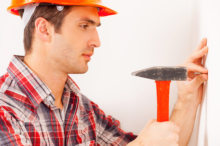 Home improvement. Side view of handsome young handyman in hardhat hammering a nail while standing near the wallの写真素材