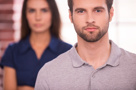 Young and successful. Confident young man looking at camera while woman standing behind him and keeping arms crossedの写真素材
