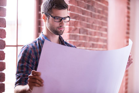 Architect examining blueprint. Confident young man in glasses examining blueprint while standing near the windowの写真素材