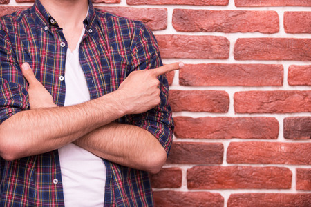 Pointing copy space. Close-up of man keeping arms crossed and pointing at copy space while standing against brick wall の写真素材