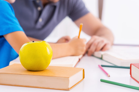 Father and son doing homework. Side view close-up image of father helping son with homework while sitting at the table together with green apple laying on the foregroundの写真素材