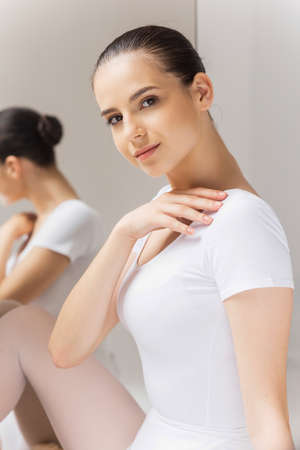 Beauty and grace. Beautiful young ballerina touching her shoulder and looking at camera while sitting near the mirrorの写真素材