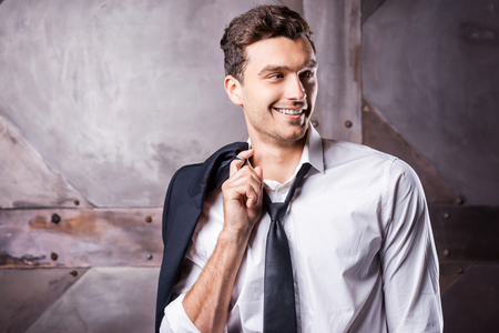 Relaxed handsome. Handsome young man in shirt and tie carrying his jacket on shoulder and looking away with smile while standing against metal backgroundの写真素材