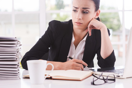 Missing the deadlines. Thoughtful young woman in suit looking at the stack of paperwork and holding head on chin while sitting at her working placeの写真素材